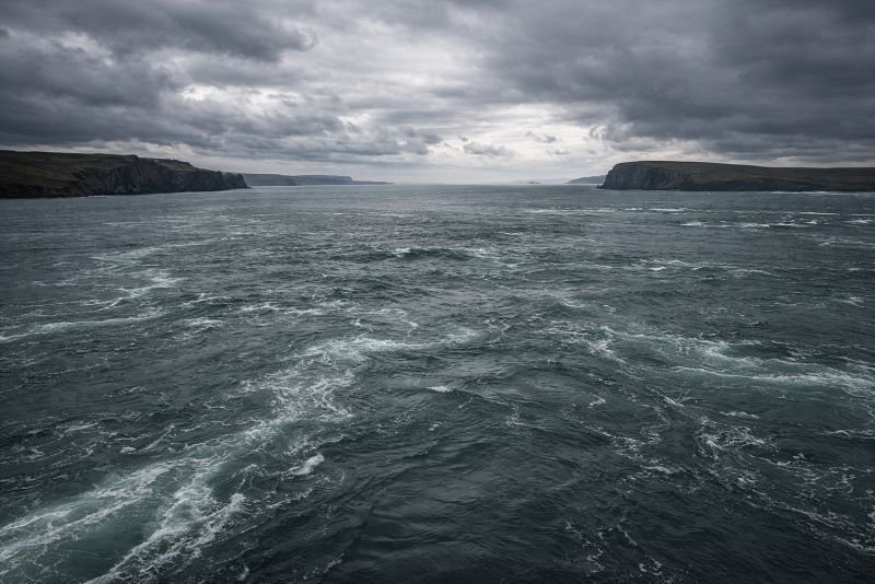 Wide view of the Pentland Firth between mainland Scotland and the Orkney Islands showing turbulent grey-green tidal currents under an overcast sky.
