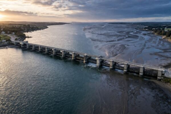 Aerial view of a tidal barrage spanning a wide estuary at low tide, showing a concrete dam with different water levels on each side under early morning light.