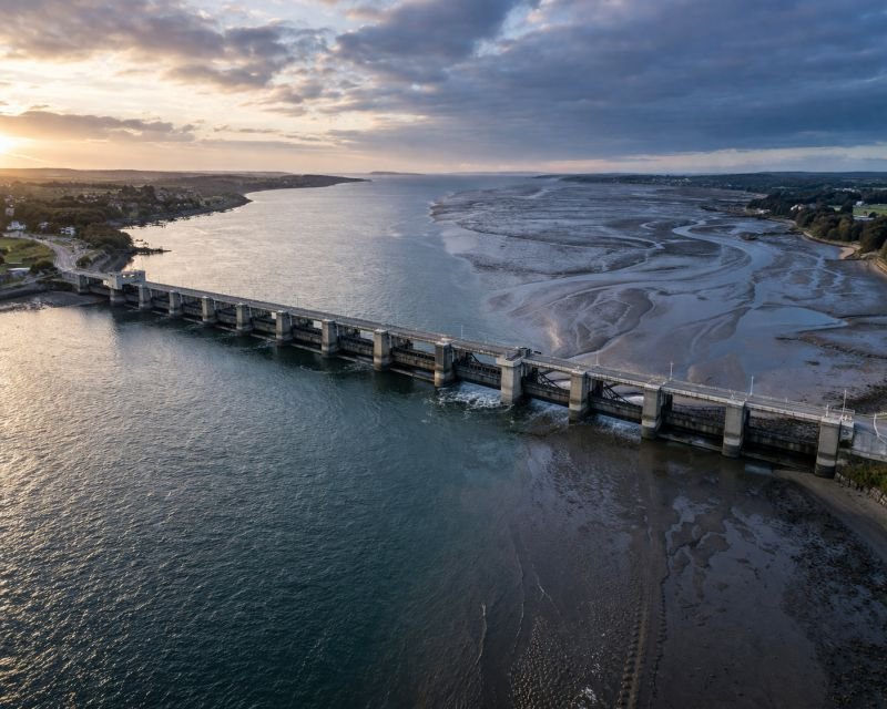 Aerial view of a tidal barrage spanning a wide estuary at low tide, showing a concrete dam with different water levels on each side under early morning light.