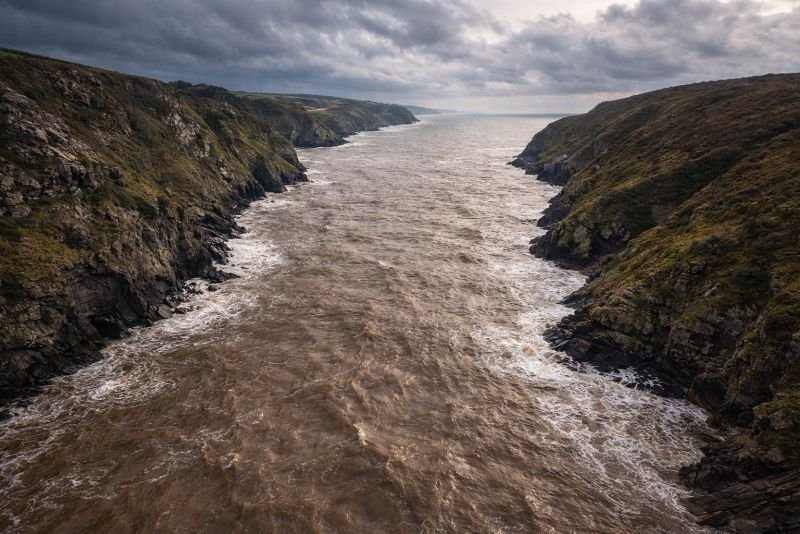 Aerial view of a narrowing estuary at high tide with turbulent brown water confined between steep rocky cliffs, illustrating tidal funnel effect.