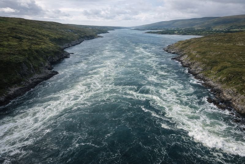 Aerial view of a narrowing estuary with strong incoming tidal flow and turbulent water, illustrating tidal resonance effects in coastal basins.