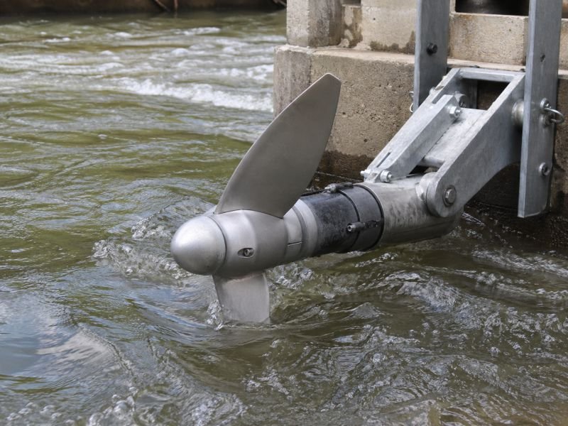 Hydrokinetic rotor with visible blade tips partially submerged in fast river water near a structural mounting frame.