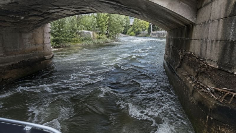 Fast-moving river current flowing beneath a stone bridge arch with debris collected along the pier base.