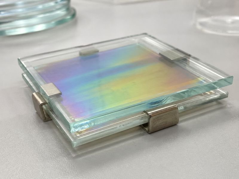 Cholesteric liquid crystal sample sealed between glass plates on a laboratory bench, showing subtle iridescent wavelength reflection under neutral light.