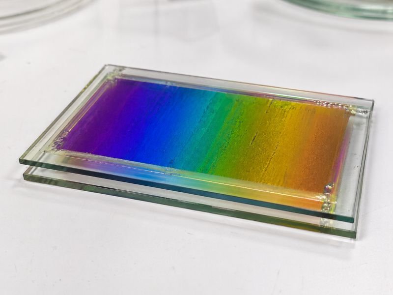 Cholesteric liquid crystal sample sealed between glass plates, showing vivid structural color banding from violet through blue and green to gold on a clean laboratory surface.