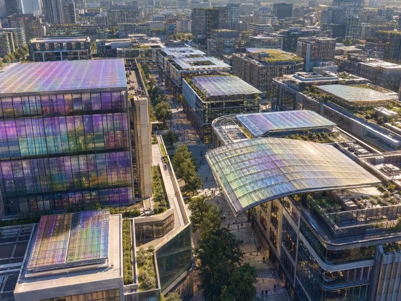 Urban district with building facades and rooftops covered in coordinated liquid crystal energy surfaces, showing subtle iridescent color variation in bright midday sunlight.