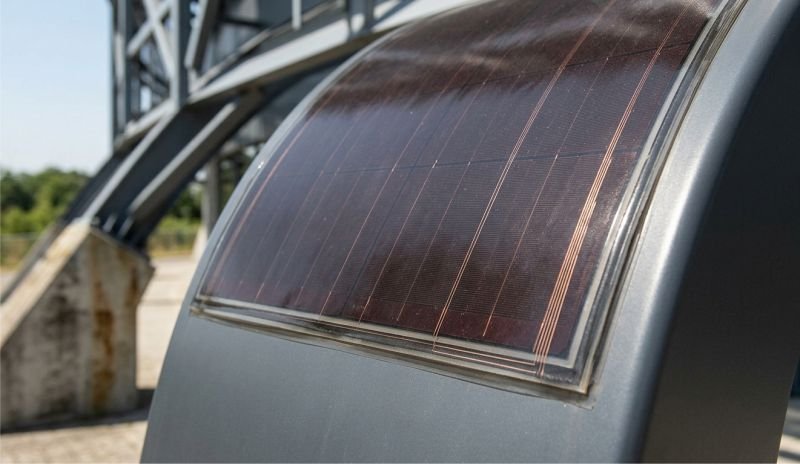 A close-up photograph of a dark, flexible thin-film photovoltaic (PV) solar panel laminated directly onto a curved gray metal structural element outdoors in daylight. The intricate grid lines and textured semiconductor surface of the thin-film solar material are visible.