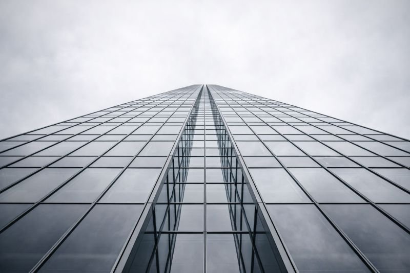 Photorealistic upward view of a futuristic glass curtain wall tower with precise geometric facade grid and converging lines, representing an active building skin in a near-future urban environment.