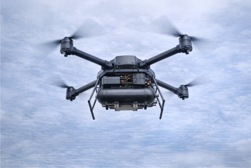 Hydrogen fuel cell drone seen from below in flight against a pale overcast sky, with compact cylindrical hardware visible beneath the airframe.
