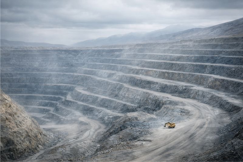 Open-pit lithium mine with grey terraced cuts and a single excavator under an overcast sky.