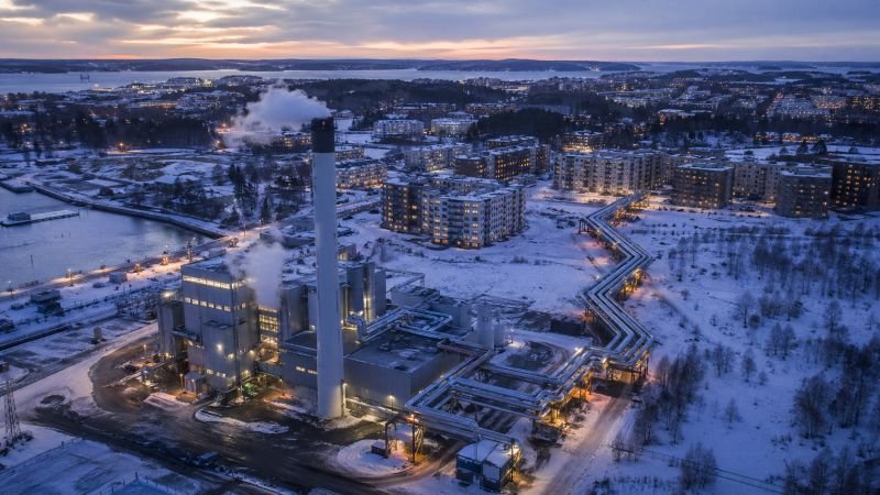 A compact combined heat and power plant with a single chimney, steam, insulated heat distribution pipes, and apartment buildings in a Scandinavian city at dusk in winter.