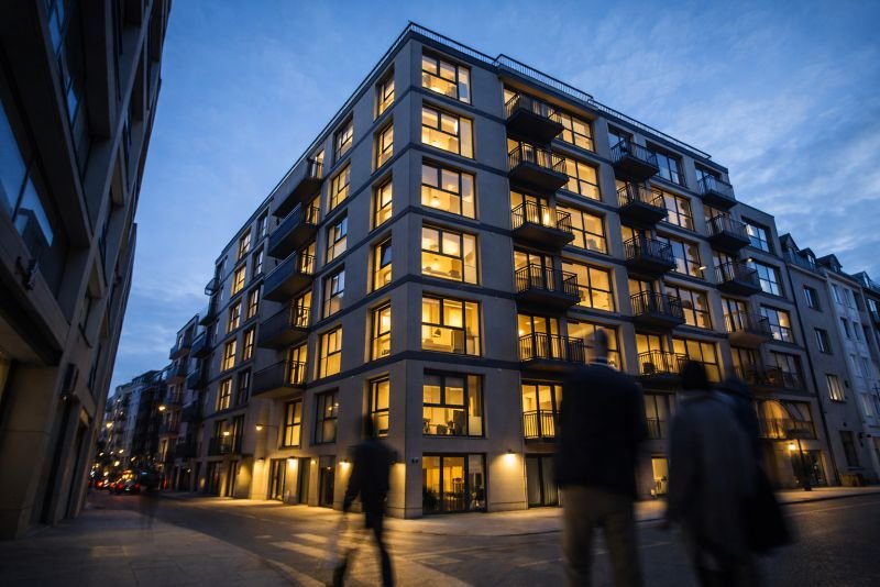 Modern mid-rise apartment building facade at dusk with warm lit windows and a narrow urban street below