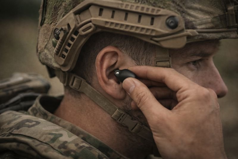 Photorealistic close-up of a tactical earpiece being fitted into a soldier’s ear in field conditions, with military helmet, camouflage uniform, and rugged outdoor environment emphasizing self-powered communication equipment.