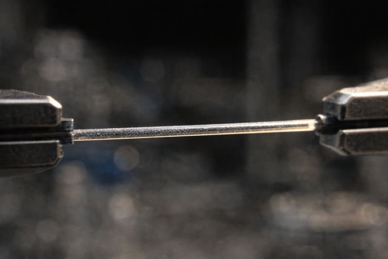 Scientific documentary-style macro photograph of a single thin photovoltaic fiber held taut between two laboratory clamps, with bright focused illumination revealing surface texture and coating layers against a dark background.