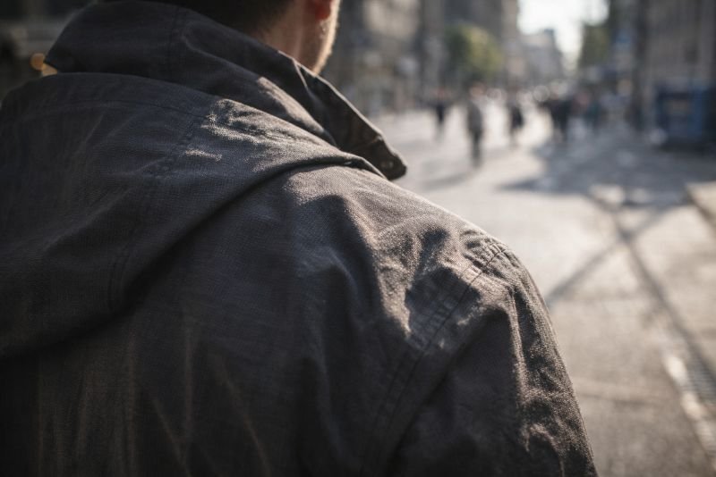 Documentary-style outdoor photograph of a person’s jacket-clad shoulder and upper back seen from behind while walking in bright direct sunlight on a city street, with sharp shadows revealing the fabric’s texture.