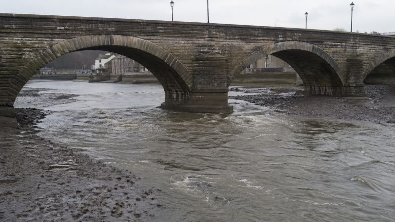 Stone arch bridge over a fast-moving tidal river with exposed mudflats and strong current beneath the arches.