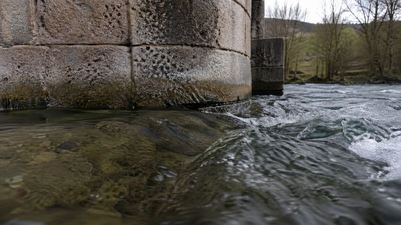 Close-up of a weathered stone bridge pier at waterline with fast river current flowing around its base.