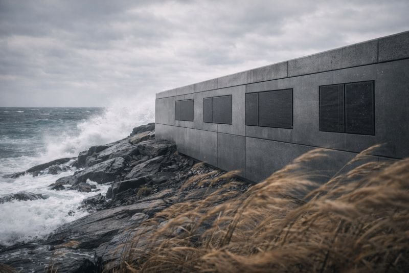 Futuristic coastal monitoring structure with integrated ultrasonic wind energy harvesting panels, set on a rocky shoreline in strong wind and rough sea under an overcast sky.