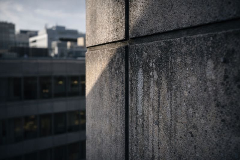 Close-up photorealistic view of a weathered concrete building facade with mineral streaking and particulate deposits, showing surface degradation under urban environmental exposure.