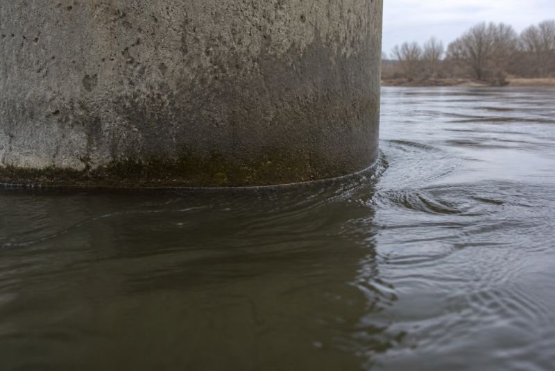 Close-up of river water flowing around a concrete bridge pier with visible surface distortion and vortex patterns downstream.
