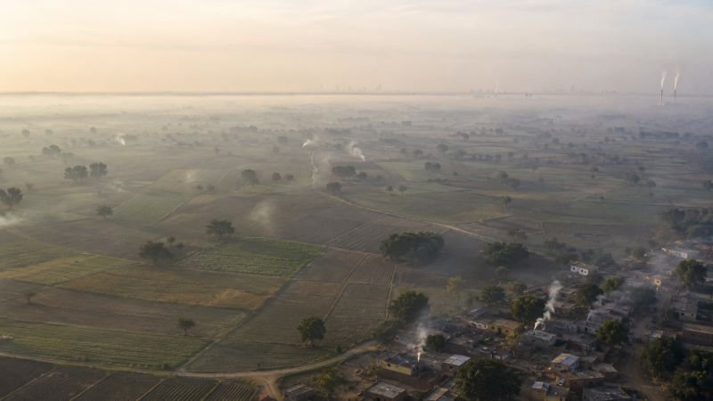 Diffuse air pollution haze over farmland and a village at dawn, with cooking fires, distant city skyline, and industrial smoke columns on the horizon