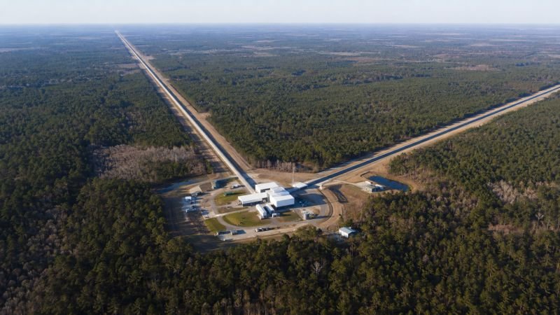 Aerial view of the LIGO Livingston detector site with two long vacuum tube arms extending through forest, showing the scale of gravitational wave research infrastructure