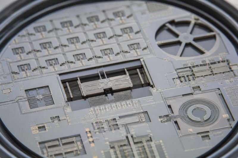 Macro view of a silicon MEMS chip with suspended microstructures, etched channels, and fine metallic traces for microsystem engineering.