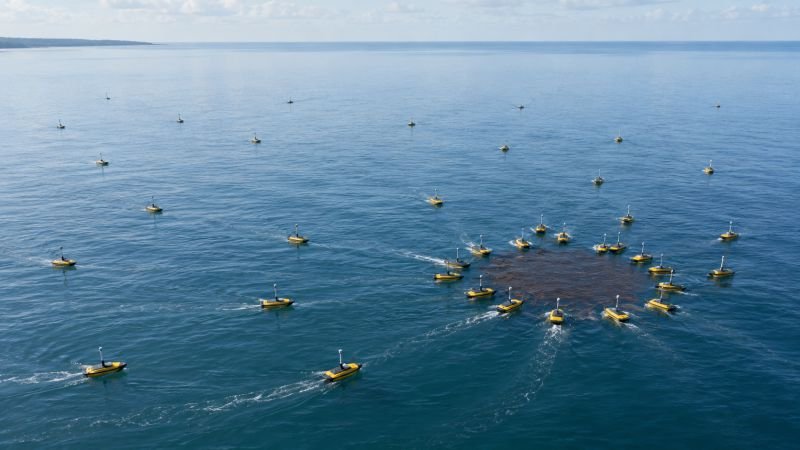 Small autonomous units spreading and regrouping across open water around a contamination zone, showing swarm intelligence in collective systems